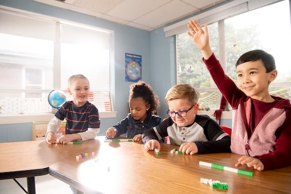 Four elementary students working together at a table in a classroom, with one student raising his hand.