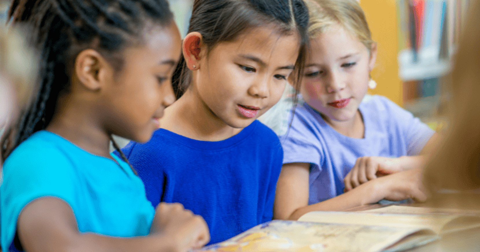 Three elementary age girls looking at a book on a desk.
